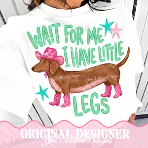 May include: A white t-shirt featuring a brown dachshund wearing a pink cowboy hat and pink cowboy boots. The text reads "WAIT FOR ME I HAVE LITTLE LEGS" in green letters, accompanied by two teal stars and one pink star above the dog illustration.