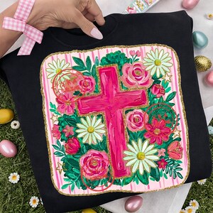 Peut inclure: Sweat-shirt noir avec une croix rose entour&eacute;e de roses roses, de marguerites blanches et de feuillage vert. La croix est centr&eacute;e sur un fond &agrave; rayures roses et blanches avec une bordure dor&eacute;e. Le motif comprend le texte "He is Risen".