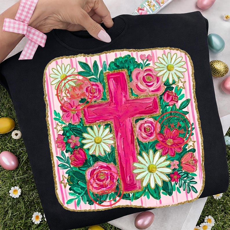 Peut inclure: Sweat-shirt noir avec une croix rose entour&eacute;e de roses roses, de marguerites blanches et de feuillage vert. La croix est centr&eacute;e sur un fond &agrave; rayures roses et blanches avec une bordure dor&eacute;e. Le motif comprend le texte "He is Risen".