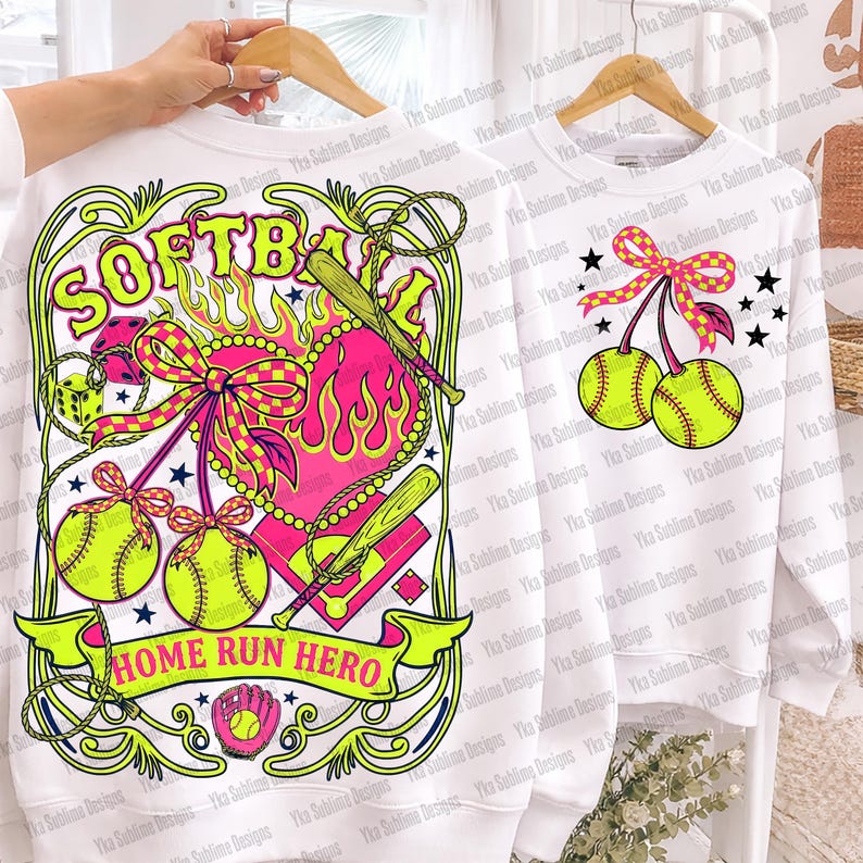 Puede incluir: Sudaderas blancas con gr&aacute;ficos de softbol. Una presenta la palabra "SOFTBALL", un coraz&oacute;n, bates de b&eacute;isbol y el texto "HOME RUN HERO". La otra tiene cerezas de softbol y estrellas.
