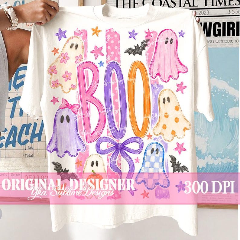 May include: A white t-shirt featuring a colorful graphic with the text BOO in large pink, purple, and orange letters. The design includes five ghosts with various patterns, small stars, two black bats, and a purple bow, set against pink stripes.