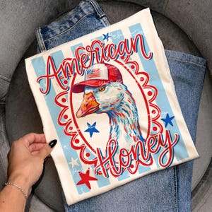 May include: Off-white t-shirt with a patriotic design featuring a goose wearing a red, white, and blue cap. The words "American Honey" are written in red script. The shirt is folded on top of blue jeans.