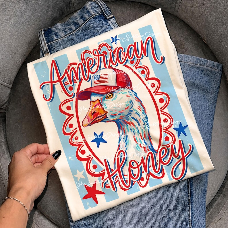 May include: Off-white t-shirt with a patriotic design featuring a goose wearing a red, white, and blue cap. The words "American Honey" are written in red script. The shirt is folded on top of blue jeans.