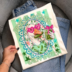 May include: Cream-colored t-shirt featuring a cartoon goose wearing heart-shaped sunglasses and holding a jar of pickles. The shirt has the text "Pickles and Goose" and "Salty & Silly" in green and teal.