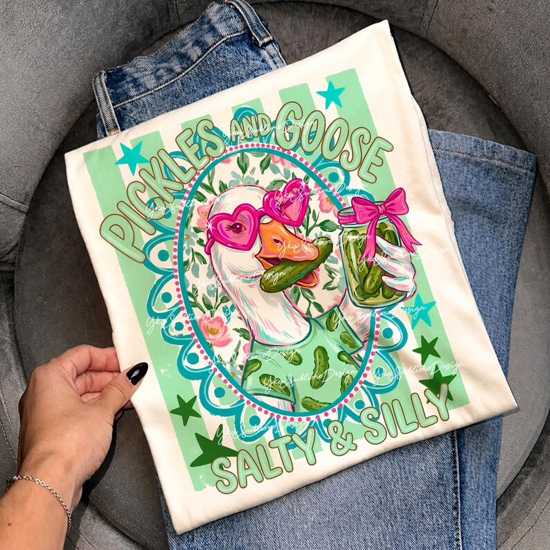 May include: Cream-colored t-shirt featuring a cartoon goose wearing heart-shaped sunglasses and holding a jar of pickles. The shirt has the text "Pickles and Goose" and "Salty & Silly" in green and teal.