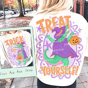 May include: A cream-coloured t-shirt featuring a graphic of a green alligator wearing a purple witch hat and cape, holding an orange jack-o'-lantern. The back displays the text "TREAT YOURSELF" in orange, while the front shows "TRICK OR".