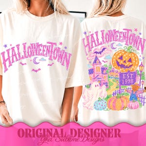 May include: An off-white t-shirt featuring a pink Halloweentown graphic on the front. The back displays a colorful illustration of a jack-o'-lantern, a ghost, a scarecrow, a house, and the text Halloweentown Est 1998 in a vibrant, sketch-like style.