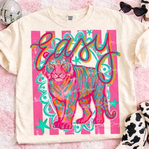 May include: Cream-colored t-shirt featuring a vibrant tiger illustration with the word "EASY" in a colorful, retro font. The tiger is depicted in shades of pink, orange, and teal, set against a pink and white striped background.