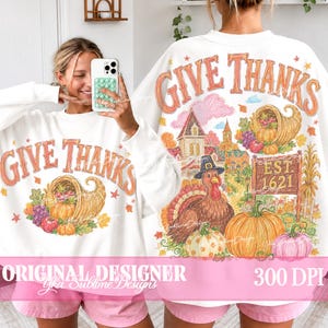 May include: A white long-sleeve sweatshirt featuring a graphic print with the text GIVE THANKS. The design includes a cornucopia filled with fruits and vegetables, a turkey wearing a pilgrim hat, a historic building, and a sign reading EST. 1621.