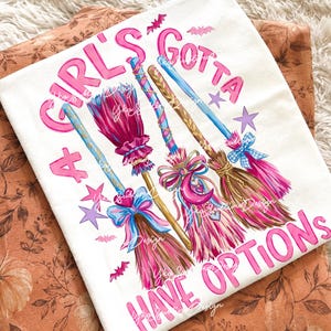 May include: A white t-shirt featuring a graphic print of three stylized broomsticks in shades of pink, blue, and brown, decorated with bows and charms. The text "A GIRL'S GOTTA HAVE OPTIONS" is printed in pink lettering, accompanied by small purple stars and pink bats.