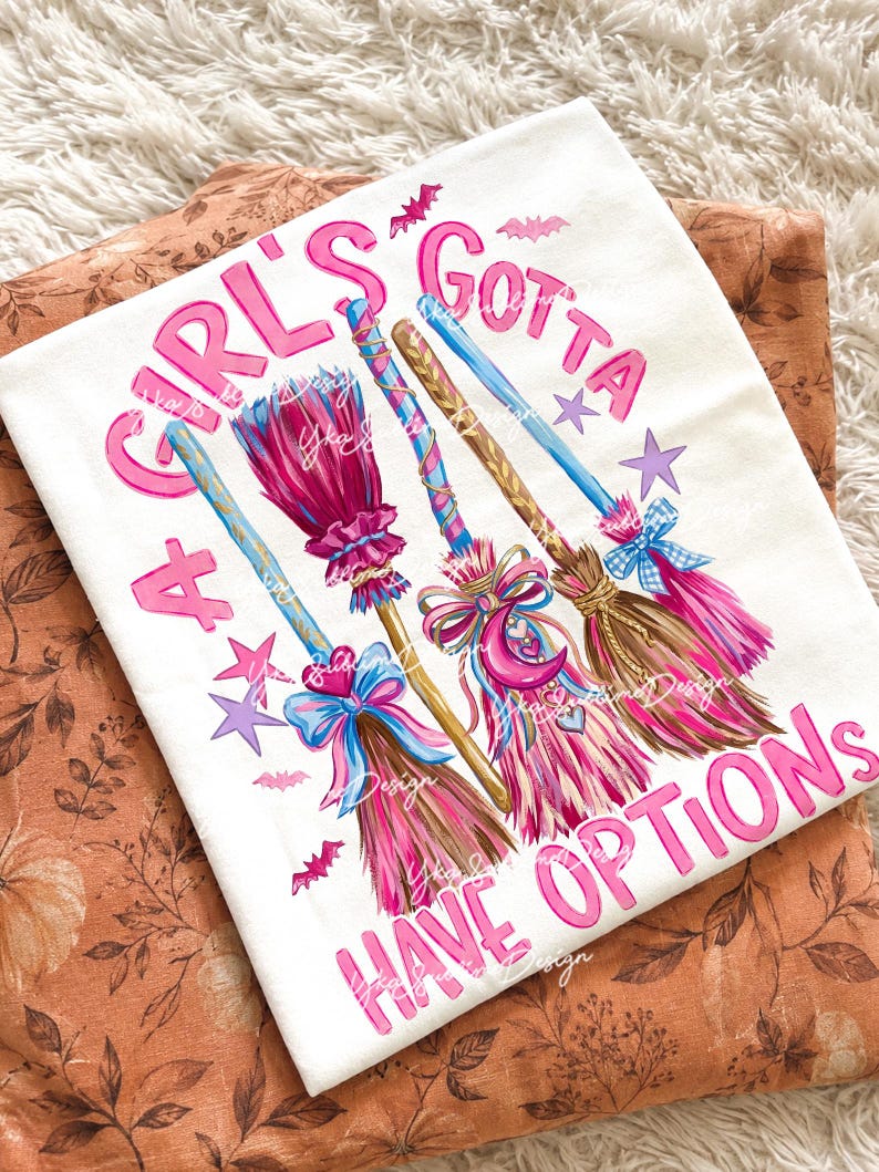 May include: A white t-shirt featuring a graphic print of three stylized broomsticks in shades of pink, blue, and brown, decorated with bows and charms. The text "A GIRL'S GOTTA HAVE OPTIONS" is printed in pink lettering, accompanied by small purple stars and pink bats.