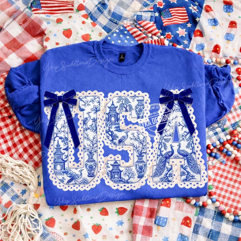 May include: A royal blue sweatshirt featuring the word "USA" in a white and blue chinoiserie pattern. The letters are edged with a scalloped trim and embellished with navy blue velvet bows. The shirt is decorated with the text "Yka Sublime Design".