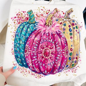 Könnte beinhalten: Ein cremefarbenes Sweatshirt mit einem Grafikdruck von drei Kürbissen. Der linke Kürbis ist blaugrün mit Schuppenmuster, der mittlere ist leuchtend pink mit Kristallverzierungen und Federn, und der rechte ist goldfarben mit bunten Edelsteinen.