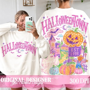 May include: A white crewneck sweatshirt featuring a pink graphic that reads HALLOWEENTOWN. The back design displays a large orange jack-o'-lantern, a ghost, a purple sign reading EST. 1998, a scarecrow, and a haunted house in vibrant colors.