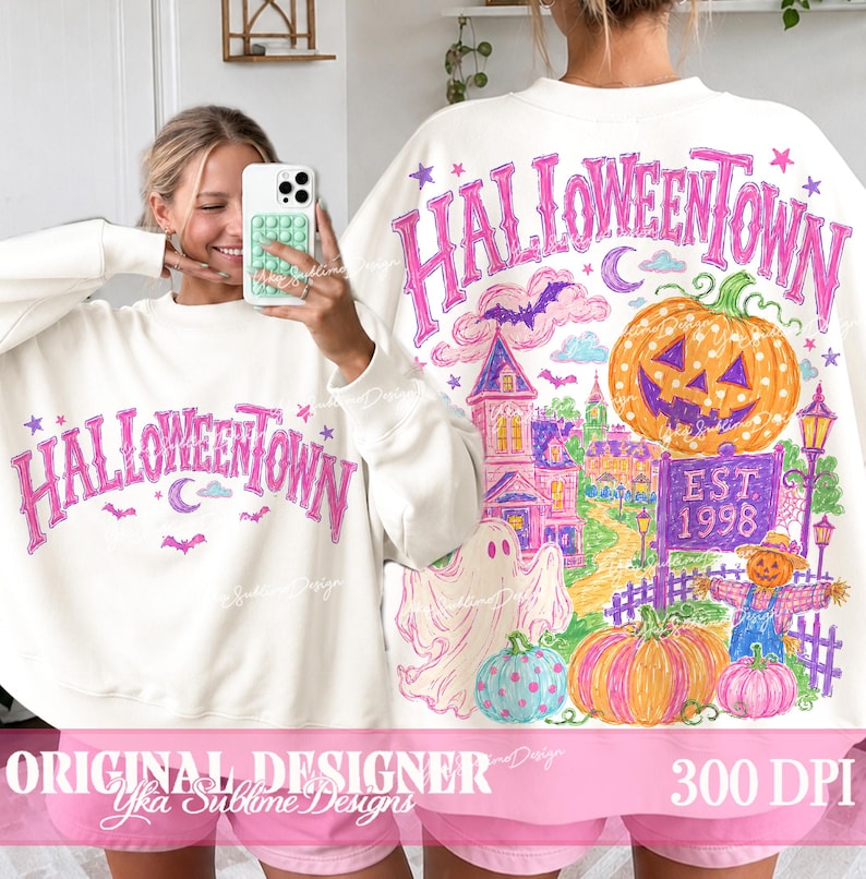 May include: A white crewneck sweatshirt featuring a pink graphic that reads HALLOWEENTOWN. The back design displays a large orange jack-o'-lantern, a ghost, a purple sign reading EST. 1998, a scarecrow, and a haunted house in vibrant colors.