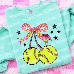 Puede incluir: Una sudadera azul claro con un dise&ntilde;o tem&aacute;tico de softbol. El gr&aacute;fico incluye dos pelotas de softbol amarillas con costuras rojas, un lazo a cuadros rosa y amarillo, estrellas negras y el texto "Yka Sublime Designs".
