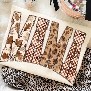 May include: A cream-colored sweatshirt featuring the word MAMA in large block letters. The letters are filled with brown and tan patterns including autumn leaves, checkered prints, small bows, acorns, and pinecones, all outlined in a scalloped border.