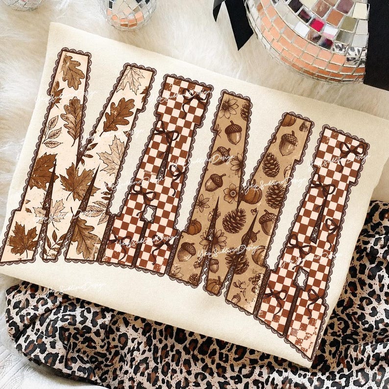May include: A cream-colored sweatshirt featuring the word MAMA in large block letters. The letters are filled with brown and tan patterns including autumn leaves, checkered prints, small bows, acorns, and pinecones, all outlined in a scalloped border.