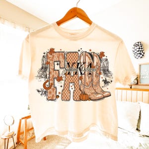May include: A cream-coloured t-shirt with a graphic design. The design features the word "FALL" in various fonts and patterns, alongside cowboy boots, autumn leaves, a horseshoe, and a pumpkin. The text "Sublime Designs" is also visible.