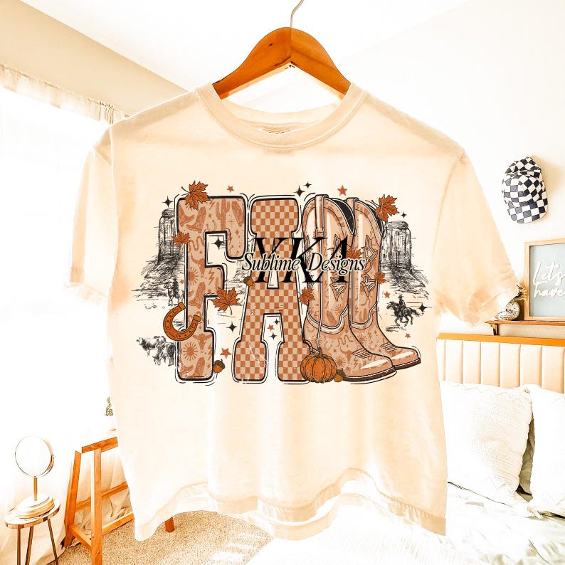 May include: A cream-coloured t-shirt with a graphic design. The design features the word "FALL" in various fonts and patterns, alongside cowboy boots, autumn leaves, a horseshoe, and a pumpkin. The text "Sublime Designs" is also visible.