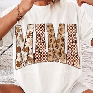 May include: A white short-sleeve t-shirt featuring the word MAMA in large block letters. The letters are filled with a pattern of brown and tan autumn leaves, a brown and white checkered design, and various fall elements like acorns and pinecones.