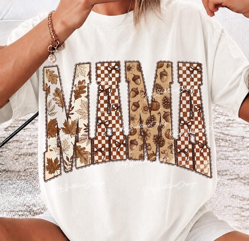 May include: A white short-sleeve t-shirt featuring the word MAMA in large block letters. The letters are filled with a pattern of brown and tan autumn leaves, a brown and white checkered design, and various fall elements like acorns and pinecones.