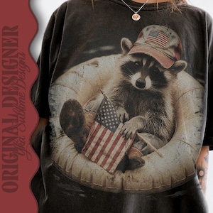 Puede incluir: Camiseta gris oscuro con un estampado desgastado de un mapache con una gorra de b&eacute;isbol con la bandera estadounidense, sentado en un flotador inflable y sosteniendo una peque&ntilde;a bandera estadounidense. La camiseta tiene el texto "ORIGINAL DESIGNER Yka Sublime Designs" en el lateral.