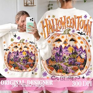 May include: A white long-sleeve sweatshirt featuring a large graphic of a jack-o'-lantern containing a spooky town scene. The text HALLOWEENTOWN is printed in orange above the pumpkin, with EST. 1998 below. The design includes bats, stars, and flowers.