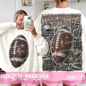 May include: A white long-sleeve sweatshirt featuring a brown football graphic on the front. The back displays a large graphic with the text TOUCHDOWN SEASON, a football, a helmet, a foam finger, a trophy, and a play diagram in a glittery style.
