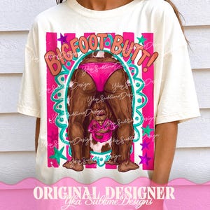 May include: Cream-colored t-shirt featuring a Bigfoot graphic. The design includes the text "BIGFOOT BUTT!" in a playful font, with a cartoon Bigfoot wearing pink swimwear. The design is framed with pink and teal accents.