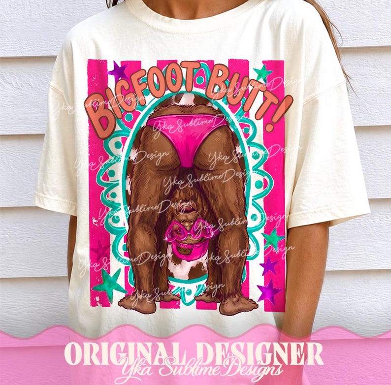 May include: Cream-colored t-shirt featuring a Bigfoot graphic. The design includes the text "BIGFOOT BUTT!" in a playful font, with a cartoon Bigfoot wearing pink swimwear. The design is framed with pink and teal accents.