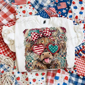 Puede incluir: Una sudadera blanca con un gr&aacute;fico de una vaca de las Tierras Altas con gafas de sol en forma de coraz&oacute;n patri&oacute;ticas y un lazo a cuadros rojo y blanco. La vaca tiene la lengua fuera. El dise&ntilde;o incorpora patrones de la bandera estadounidense y estrellas. El texto "S FRE OM" es visible.
