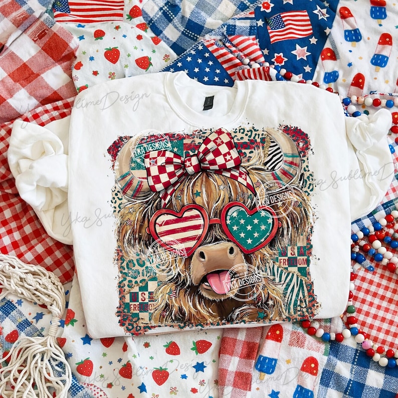 Puede incluir: Una sudadera blanca con un gr&aacute;fico de una vaca de las Tierras Altas con gafas de sol en forma de coraz&oacute;n patri&oacute;ticas y un lazo a cuadros rojo y blanco. La vaca tiene la lengua fuera. El dise&ntilde;o incorpora patrones de la bandera estadounidense y estrellas. El texto "S FRE OM" es visible.