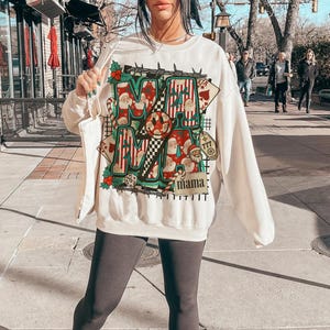 May include: Cream-coloured sweatshirt with a festive Christmas-themed graphic. The design features the word "MAMA" in a bold, colourful font, with Santa Claus illustrations and playing card motifs. The sweatshirt has long sleeves.
