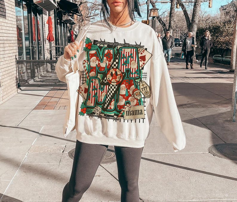 May include: Cream-coloured sweatshirt with a festive Christmas-themed graphic. The design features the word "MAMA" in a bold, colourful font, with Santa Claus illustrations and playing card motifs. The sweatshirt has long sleeves.