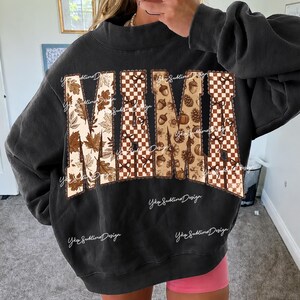 May include: A charcoal gray crewneck sweatshirt featuring the word MAMA in large block letters. The letters are filled with a pattern of brown autumn leaves, a brown and white checkerboard print, and small brown acorns and pinecones.
