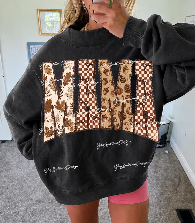 May include: A charcoal gray crewneck sweatshirt featuring the word MAMA in large block letters. The letters are filled with a pattern of brown autumn leaves, a brown and white checkerboard print, and small brown acorns and pinecones.