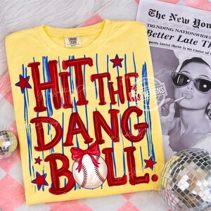 Puede incluir: Camiseta amarilla con la frase "HIT THE DANG BALL" en letras rojas, rayas verticales azules y estrellas rojas. Una pelota de b&eacute;isbol con un lazo rojo aparece debajo del texto. La camiseta se muestra sobre una superficie estampada.
