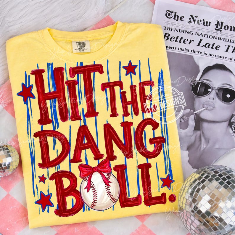 Puede incluir: Camiseta amarilla con la frase "HIT THE DANG BALL" en letras rojas, rayas verticales azules y estrellas rojas. Una pelota de b&eacute;isbol con un lazo rojo aparece debajo del texto. La camiseta se muestra sobre una superficie estampada.