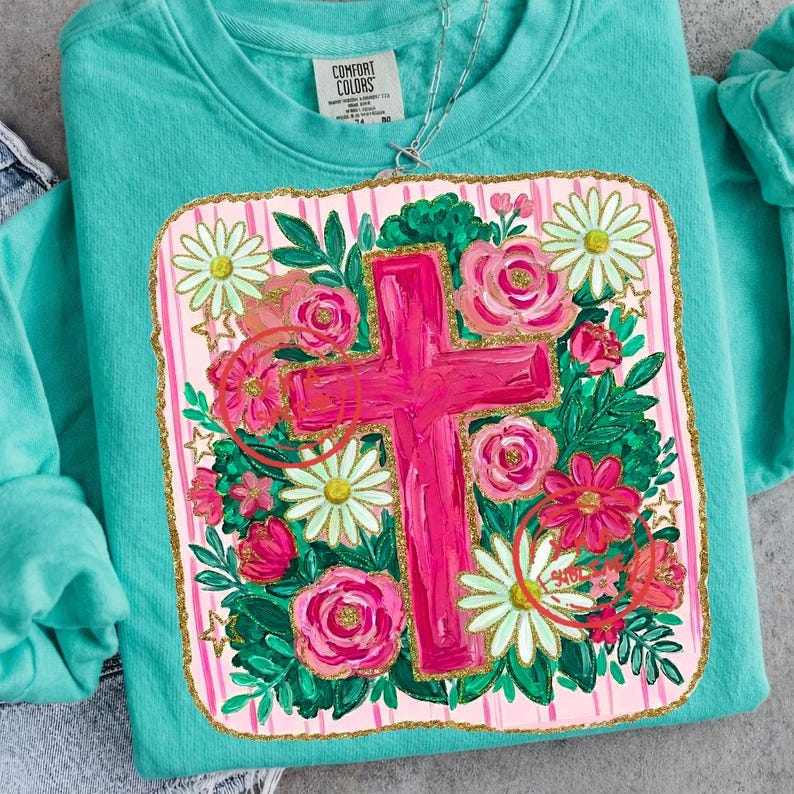 Peut inclure: Un sweat-shirt Comfort Colors turquoise avec un motif de croix rose entour&eacute; de fleurs. La croix est le point central, avec des roses roses, des marguerites et des feuilles vertes. Le motif a une bordure dor&eacute;e scintillante.