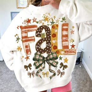 Puede incluir: Una sudadera de cuello redondo color crema con la palabra FALL en letras grandes y decorativas. Las letras tienen rayas naranjas, lunares blancos sobre fondo marrón y pequeñas calabazas naranjas. Debajo hay un gran lazo de cuadros verde.