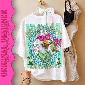 May include: White t-shirt with a whimsical graphic of a goose wearing heart-shaped sunglasses and holding a jar of pickles. The design includes the text "Pickles and Goose" and "Salty & Silly" in a playful font. The shirt is hanging on a white hanger.