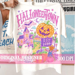 May include: A white short-sleeve t-shirt featuring a colorful graphic print with the text HALLOWEENTOWN and EST. 1998. The design includes a large orange jack-o'-lantern, a pink haunted house, a white ghost, a scarecrow, and purple bats.