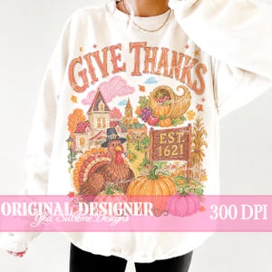 May include: A cream-colored crewneck sweatshirt featuring a colorful graphic print with the text GIVE THANKS and EST 1621. The design includes a turkey wearing a hat, a large orange pumpkin, a cornucopia, a house, and autumn foliage.