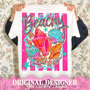 Op de afbeelding: Wit t-shirt met een strandthema. Het ontwerp toont het woord "Beachy" in roze en goud, samen met kleurrijke schelpen in tinten roze, oranje en turquoise. Roze en witte strepen vormen de achtergrond.