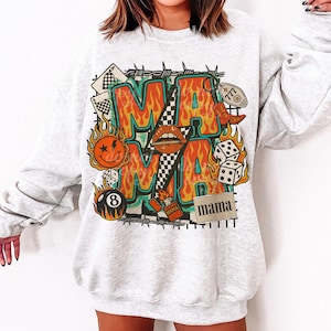 Puede incluir: Una sudadera gris claro con un dise&ntilde;o gr&aacute;fico colorido. El dise&ntilde;o presenta la palabra "MAMA" en letras grandes con forma de llama, rodeada de varias ilustraciones, incluyendo dados, una carita sonriente y una bota de vaquero.