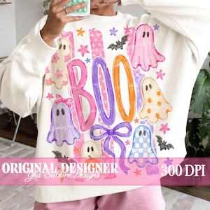 May include: A cream-colored long-sleeve sweatshirt featuring a colorful graphic print with the word BOO in large pink, purple, and orange letters. The design includes various ghosts, bats, stars, and bows in pink, purple, and orange patterns.