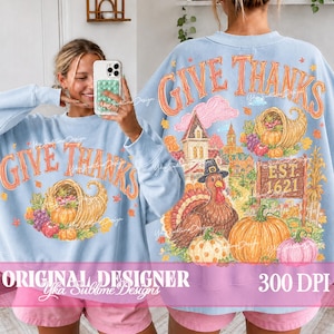 May include: A light blue long-sleeve sweatshirt featuring a graphic print with the text GIVE THANKS. The design includes a cornucopia, a turkey wearing a hat, a house, and pumpkins. The back of the sweatshirt displays the same detailed illustration.