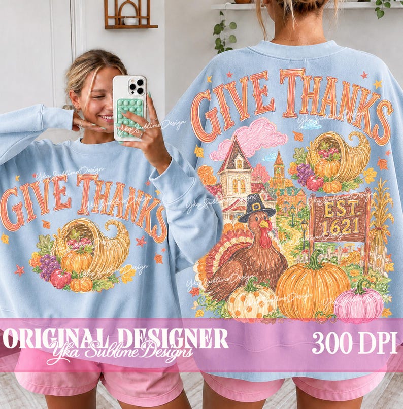 May include: A light blue long-sleeve sweatshirt featuring a graphic print with the text GIVE THANKS. The design includes a cornucopia, a turkey wearing a hat, a house, and pumpkins. The back of the sweatshirt displays the same detailed illustration.