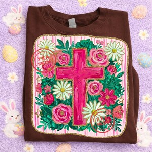 Peut inclure: Un sweat-shirt marron avec une croix rose entour&eacute;e de roses roses, de marguerites blanches et de feuilles vertes. Le motif est pos&eacute; sur un fond &agrave; rayures roses et blanches avec une bordure dor&eacute;e. Le sweat-shirt est pli&eacute; sur une surface violette.